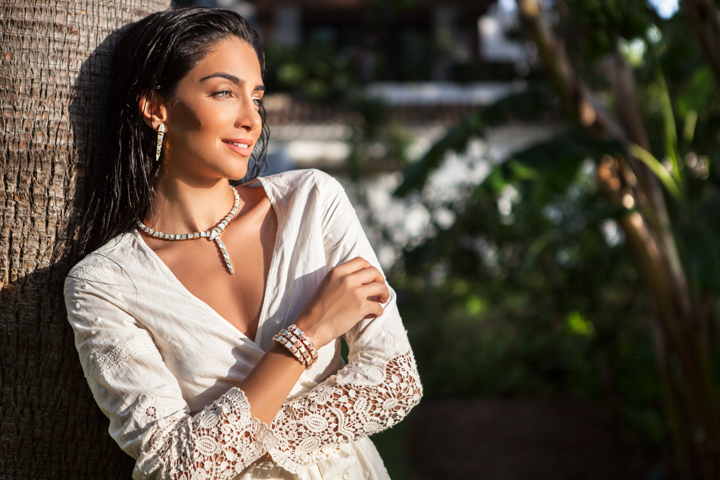 A woman wearing layered necklaces rests against a tree in a garden.
