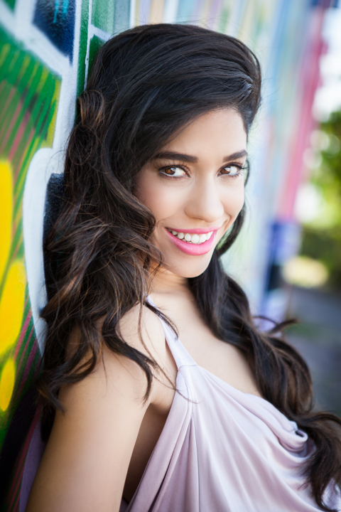 A woman with long dark curls looks toward the camera beside a colorful wall.