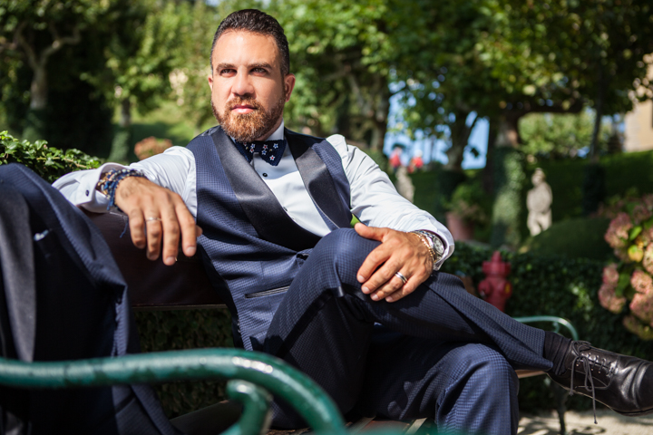 A man wearing a bow tie sits on a park bench surrounded by trees.