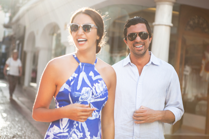 Two laughing adults wearing sunglasses stand together on a sunny street.