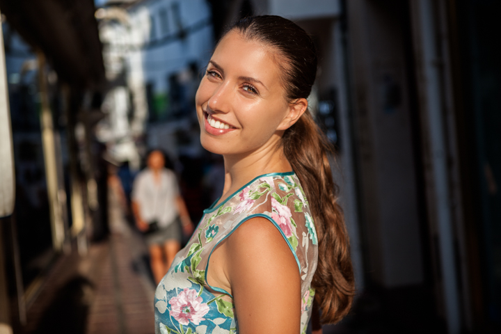 A smiling woman in a floral top looks back on a city street.