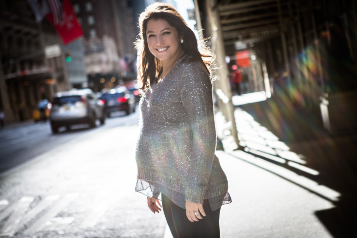 A smiling woman stands beneath a covered walkway on a sunny street.