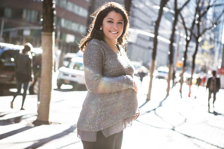 A smiling woman stands on a tree-lined city sidewalk in bright sunlight.