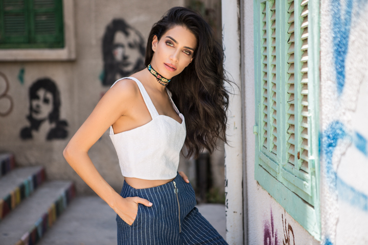 A woman in a white top and blue trousers stands beside painted shutters.