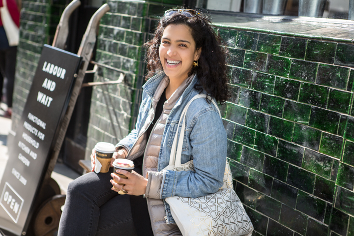 A smiling woman holds a takeaway cup beside a dark green tiled wall.