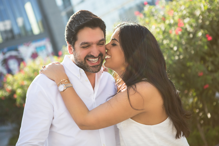 A woman kisses a smiling man's cheek in a sunny garden.