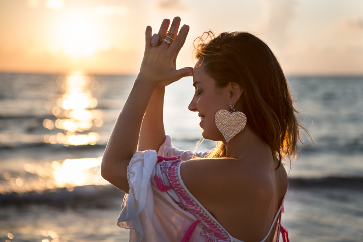 A woman raises a hand to her forehead beside the sea at sunset.