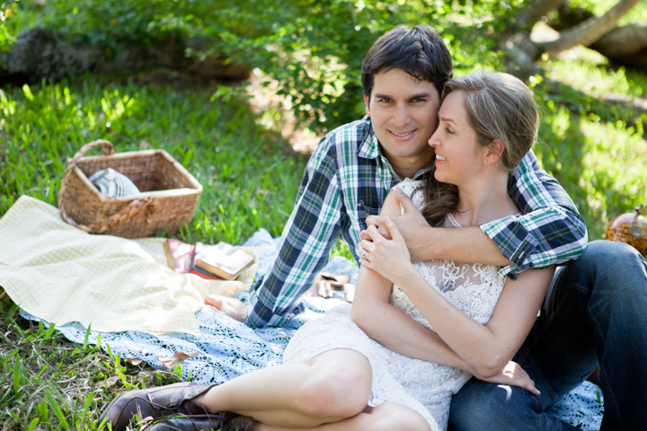 Two adults cuddle on a picnic blanket beneath garden trees.