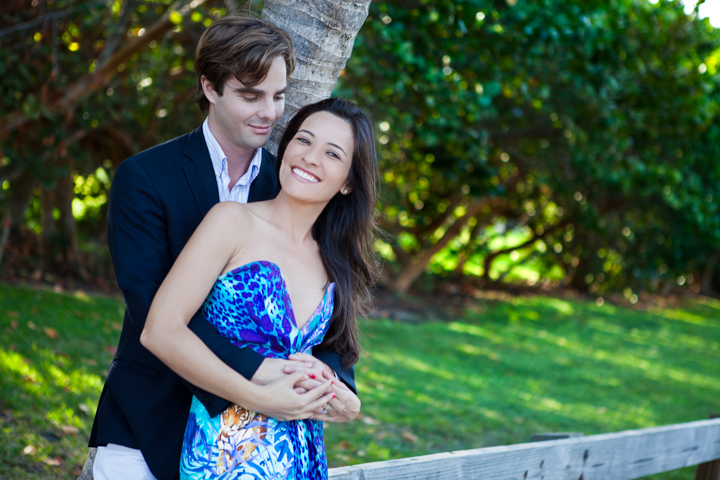 A man embraces a woman in a patterned blue dress beneath green trees.