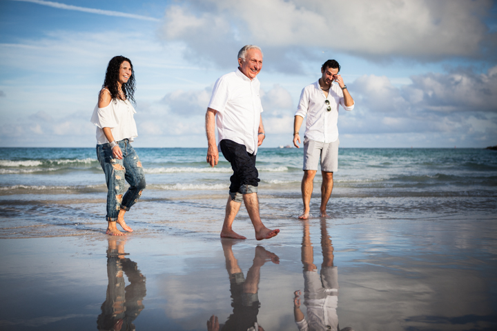 Three adults walk through shallow ocean water beneath a cloudy sky.
