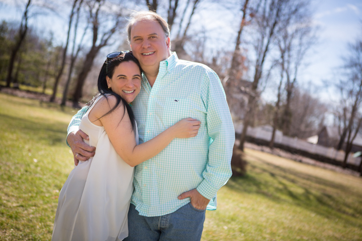 Two adults cuddle beneath bare trees on a sunny lawn.