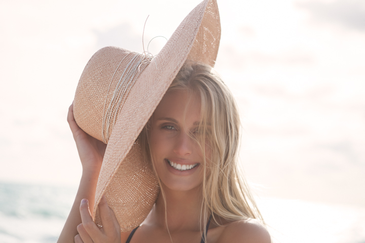 A smiling woman tips a wide-brimmed straw hat toward the camera.