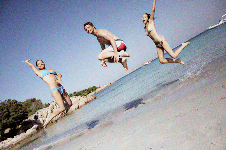 Three adults leap above wet sand at the edge of the ocean.