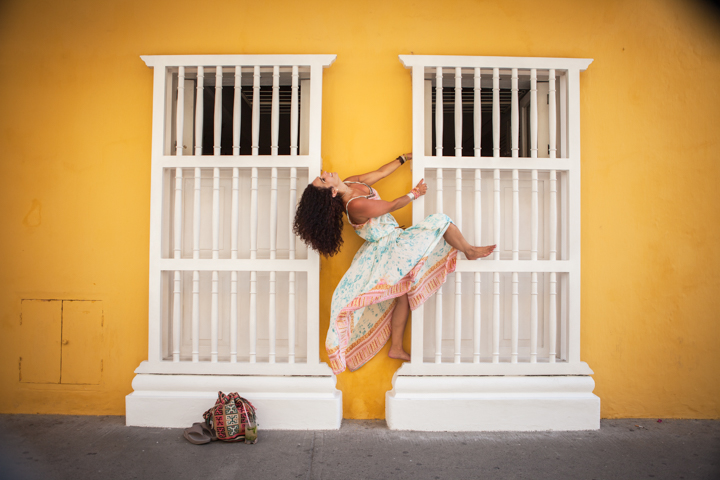 A person balances on one leg against a yellow wall with white barred windows.