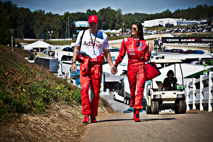 Two adults in red racing suits walk hand in hand near parked vehicles.