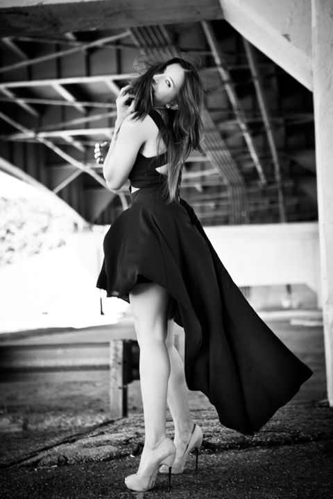 A woman in a long black dress stands beneath a staircase in black and white.