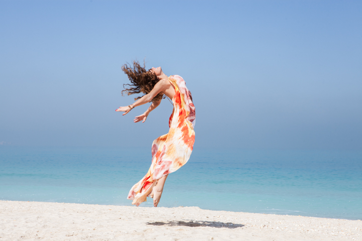 A woman in a flowing colorful dress jumps beside turquoise ocean water.