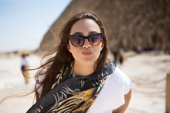 A woman in dark sunglasses and a patterned scarf stands on a sandy beach.