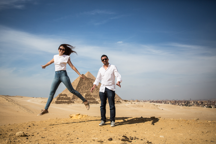 Two adults jump together in a wide sandy landscape beneath a blue sky.