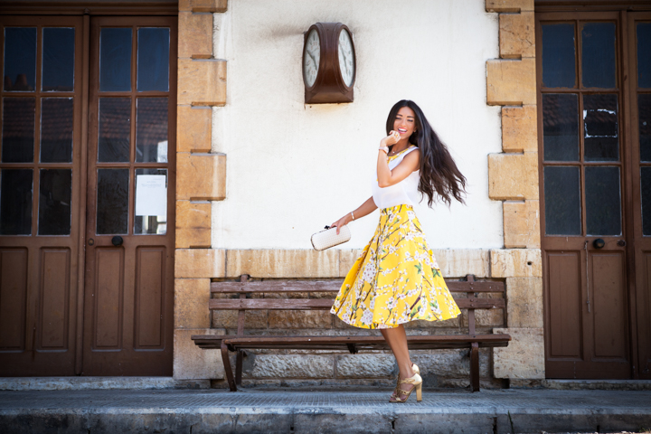 A woman in a yellow skirt stands beside a wooden bench and doorway.