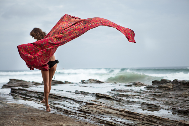A person holds flowing red fabric above coastal rocks beside the sea.