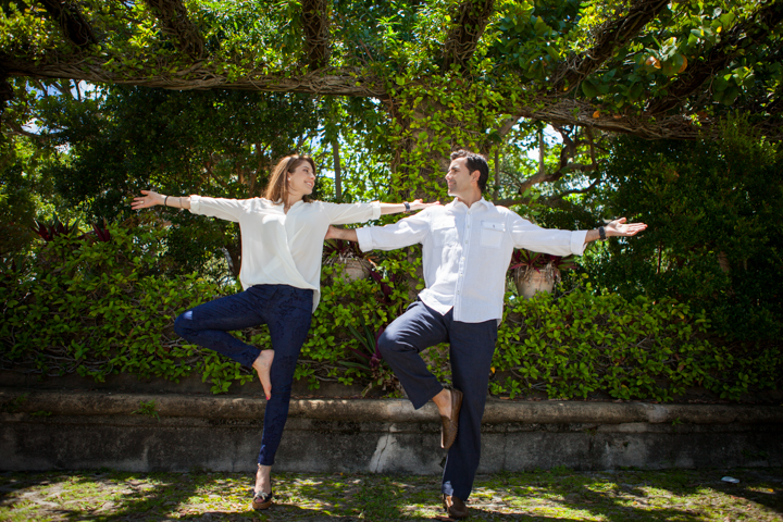 Two adults balance on one leg with arms spread in a garden.