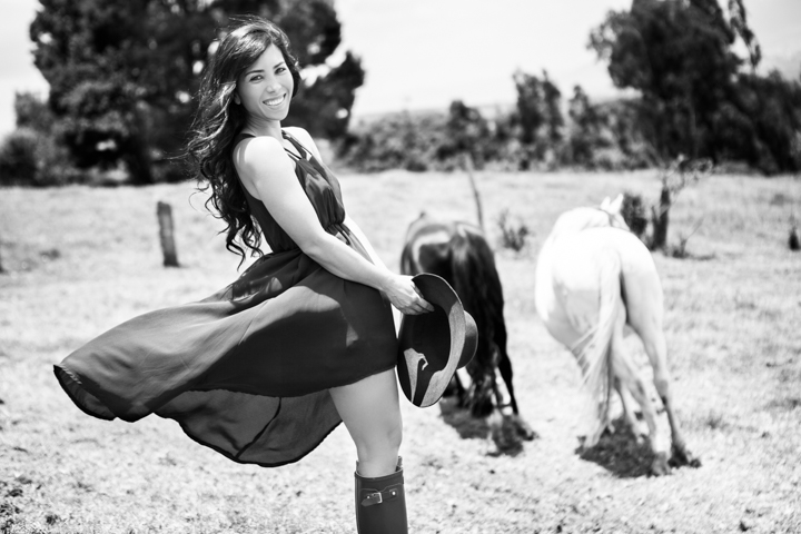 A woman turns in a dark dress beside grazing horses in black and white.