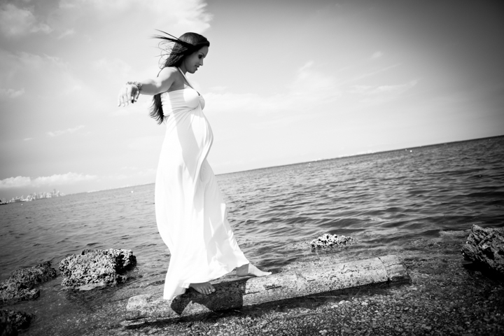 A woman in a long white dress stands beside the sea in a black-and-white photograph.
