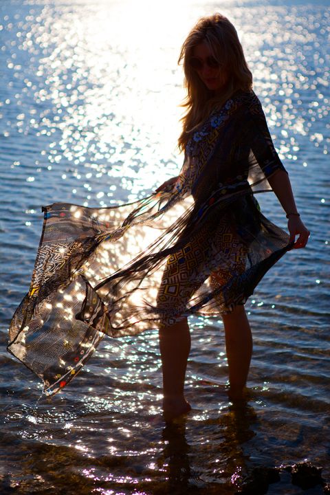 A person stands in shallow water holding flowing fabric against the sparkling sea.