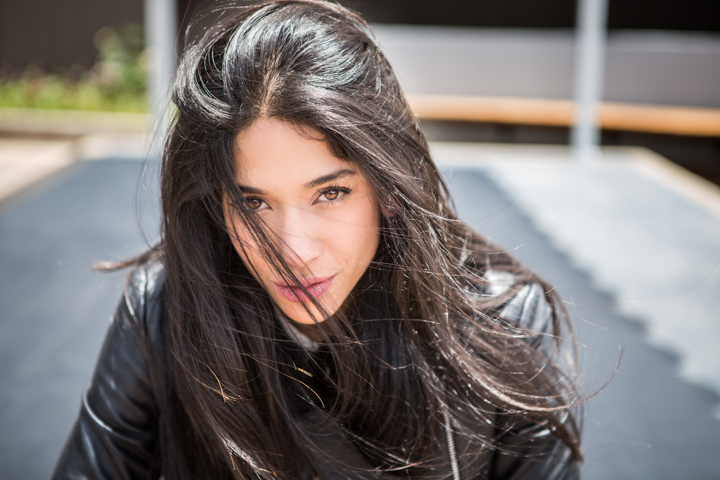 A woman with long dark hair wears a black jacket in bright outdoor light.