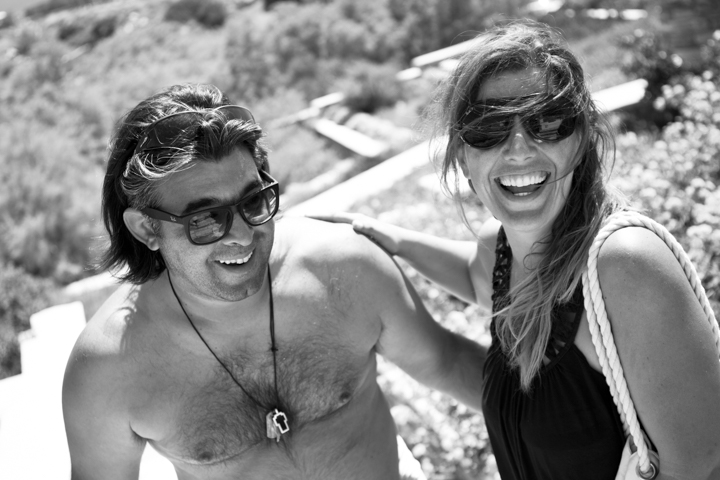 Two adults wearing sunglasses laugh together in a black-and-white portrait.