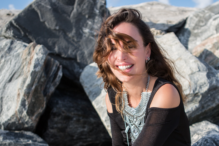 A smiling woman sits among coastal rocks with the wind blowing her hair.