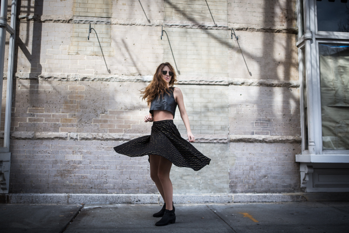 A woman turns in a black skirt beside a pale city wall.