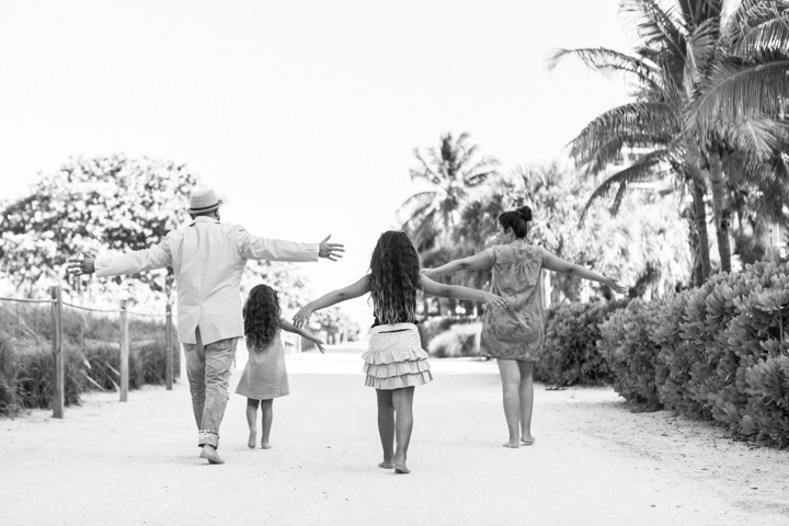 A woman and two children walk hand in hand along a palm-lined path in black and white.