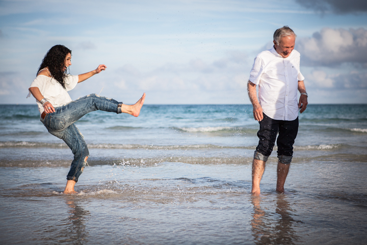 Two adults play in shallow ocean water, one lifting a leg toward the waves.