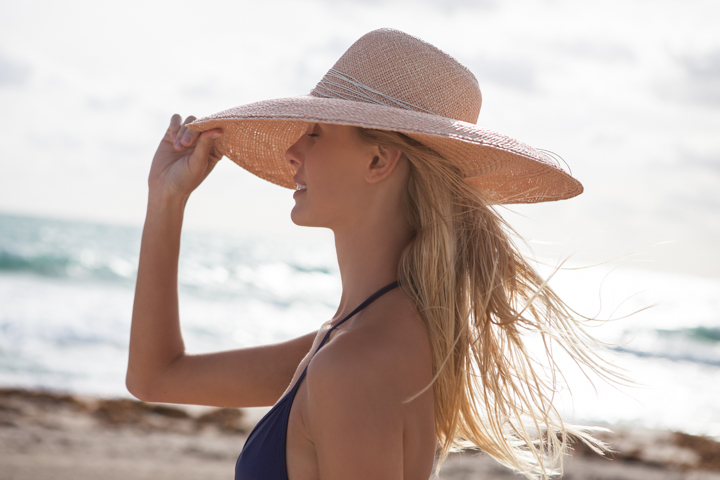A woman holds the brim of a wide pink hat beside the ocean.