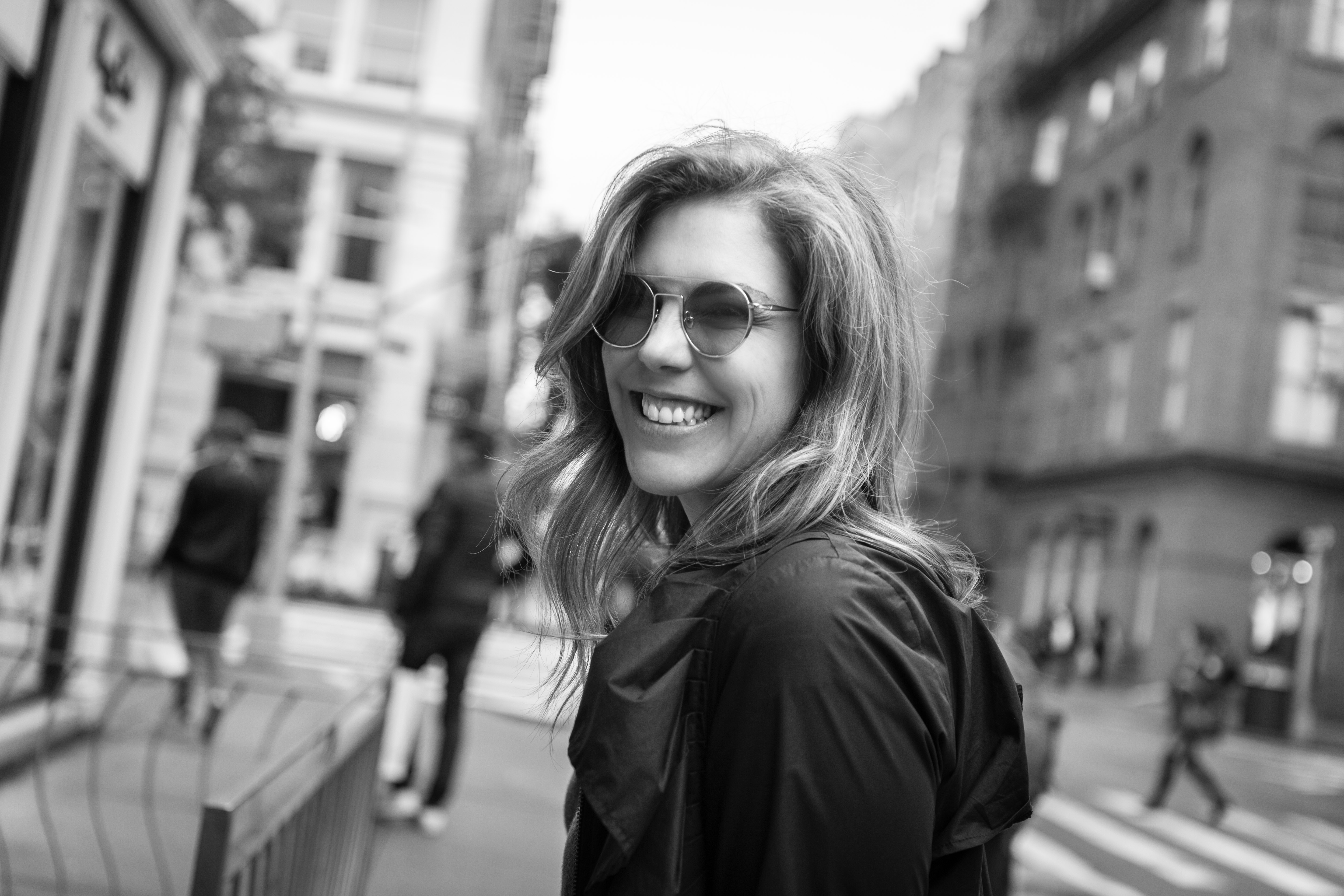 A woman wearing sunglasses looks back toward the camera on a city street.