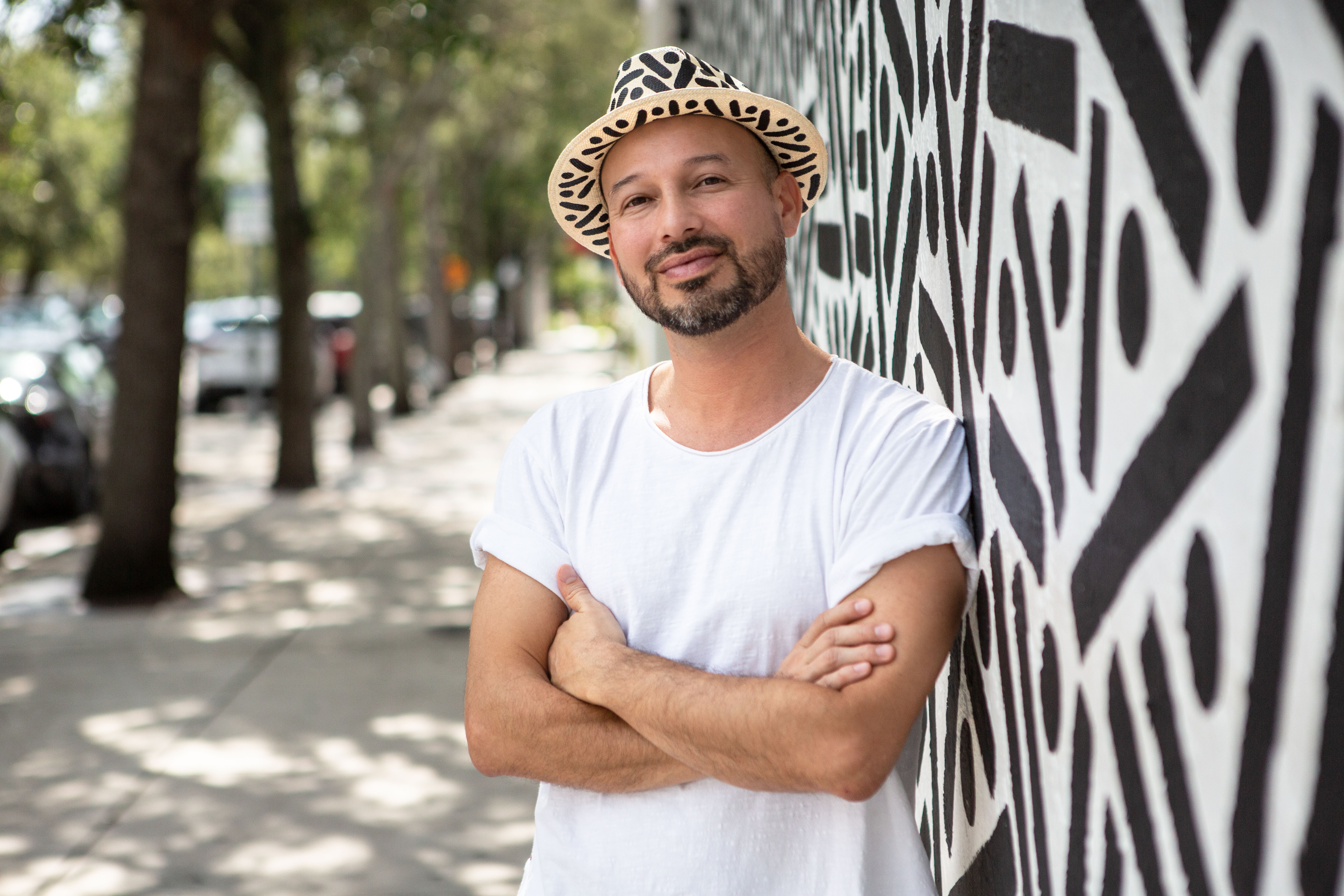 Pipe Yanguas stands beside a black-and-white mural wearing a white shirt and patterned hat.