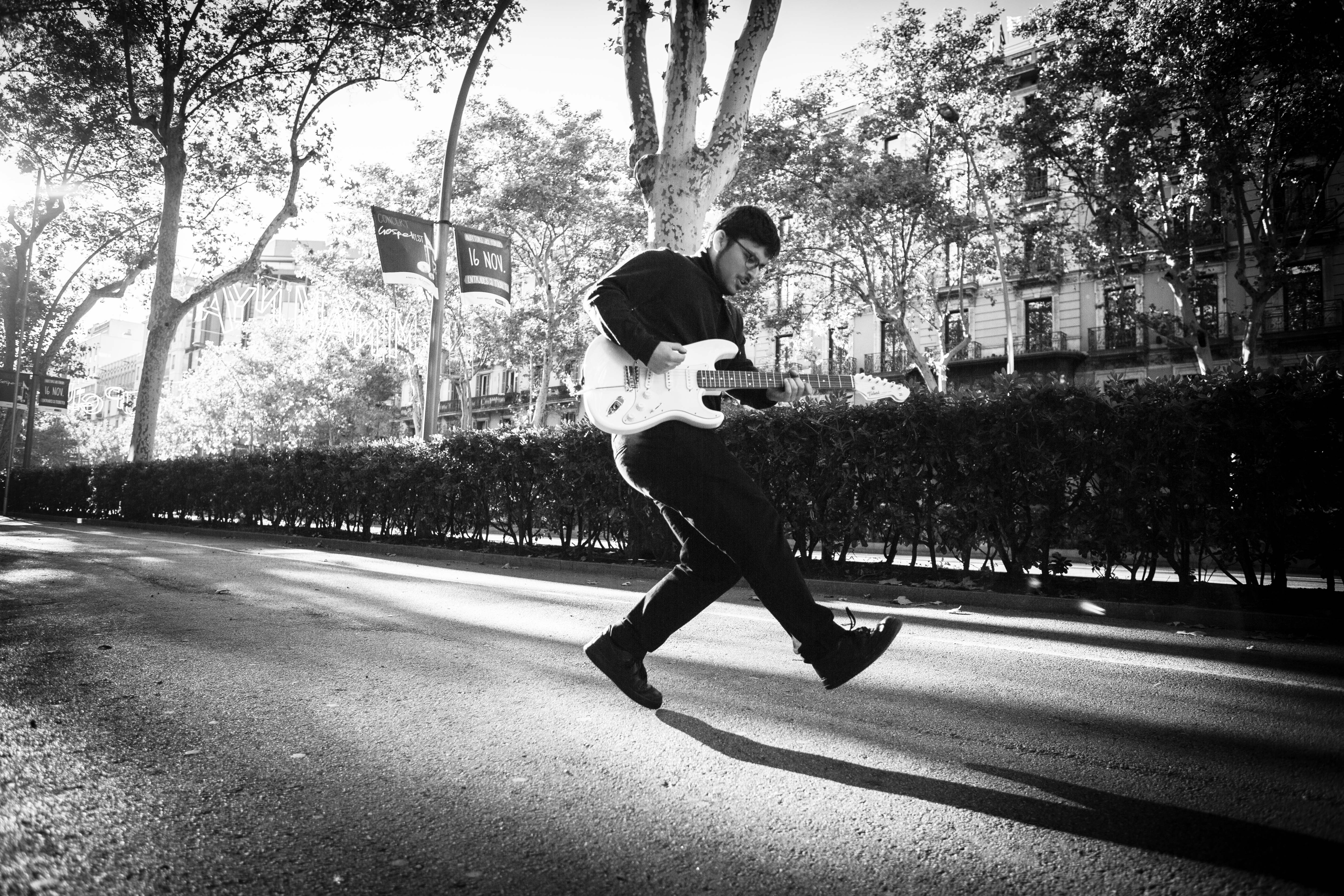 A man leaps across a tree-lined street with an electric guitar in a black-and-white photograph.